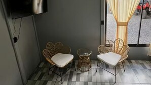 Quarto com televisão na parede, duas cadeiras de vime com almofadas, piso estampado e ventilador de teto.