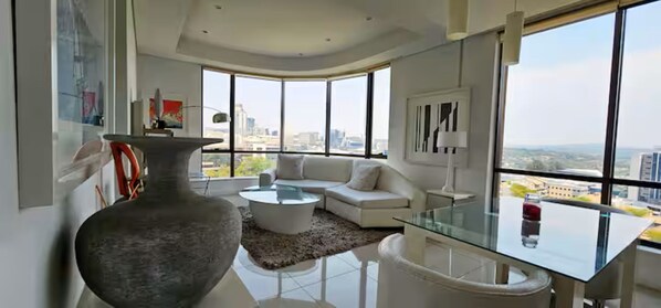 Un salon moderne avec une table basse en verre, un canapé blanc, un grand vase et une vue sur la ville à travers de grandes fenêtres.