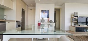 Une cuisine moderne avec une table à manger en verre, des appareils électroménagers intégrés et un téléviseur dans l’espace de vie.