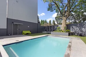 En swimmingpool med tilhørende terrasse, et træ og en moderne bygning.