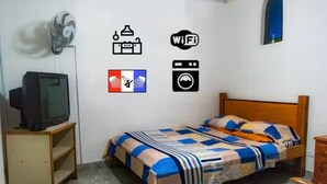Et soveværelse med en seng, et fjernsyn på et stativ, en ventilator samt vægikoner, der angiver faciliteter som WiFi, vaskeri og TV.