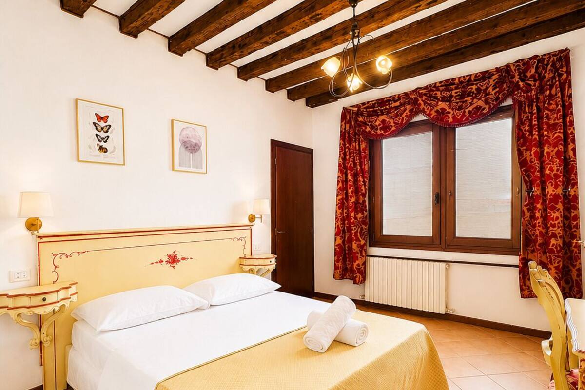 A bedroom with a wooden ceiling, a bed with a yellow bedspread, two framed pictures on the wall, a window with red curtains, and a radiator.