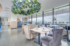 Ein modernes Restaurant mit hohen Decken, großen Fenstern und einer zentral platzierten, hängenden Blumenkomposition.