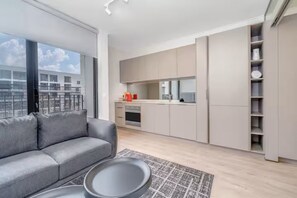Eine moderne Wohnung mit grauem Sofa, einem Esstisch, einer Küche mit grauen Schränken und Blick auf Gebäude durch große Fenster.