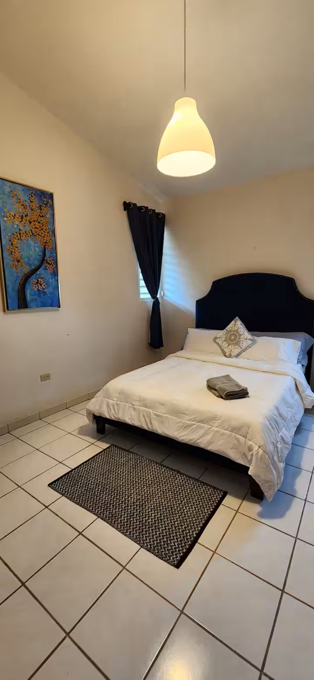 A bedroom with a bed, a hanging light fixture, a painting on the wall, and a window with curtains.