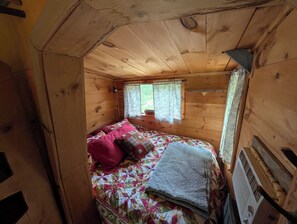 A small, cozy cabin room with a wooden interior, a bed with a colorful quilt, a window with curtains, and a heater. - Tiny House Oasis in beautiful Waldo County, Maine. Dogs welcome! JUNE PRICE DROP (Jackson)
