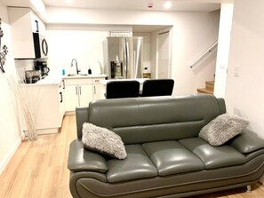 Een moderne woonkamer met een grijze leren bank, twee zachte kussens, en een keuken in de achtergrond met witte kastjes, een roestvrijstalen koelkast en een koffiezetapparaat.
