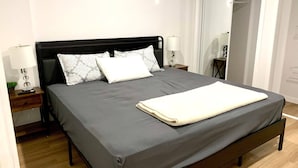 A neatly made bed with a grey bedspread and white pillows, a bedside table with a lamp, and a mirror on the wall. - Royal Oak- 2-bedroom Suite (Winnipeg)