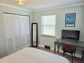 Ein Schlafzimmer mit einem Bett, einem Schreibtisch, einem Sessel, einem Fernseher, einem Fenster mit Jalousien, einem Spiegel und einer Tür mit dekorativen Paneelen.