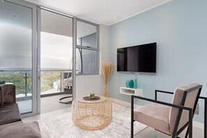 Ein modernes Wohnzimmer mit großem Fenster, Flachbildfernseher, rundem Rattan-Tisch und einer Couch.