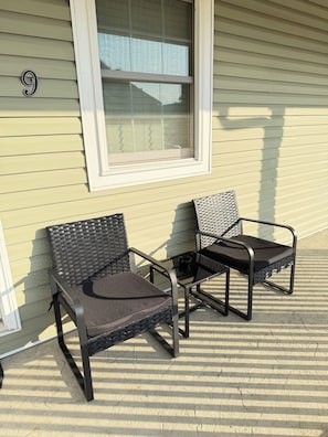 Two outdoor patio chairs with metal frames and woven seats, positioned on a textured patio surface, next to a building with a window and a number '9' sign.