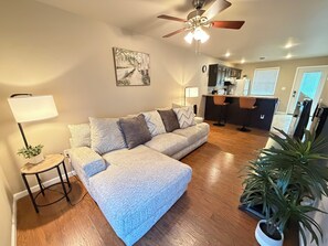A living room with a sofa, a lamp, a ceiling fan, a painting, and a kitchen area.