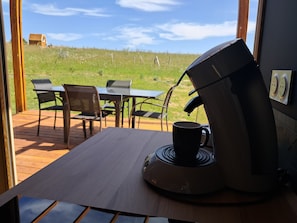A coffee machine on a table with a cup, a grill outside, and a view of a grassy field.