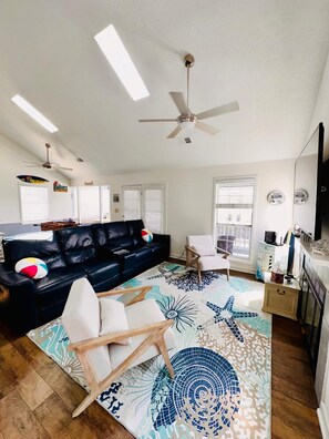 A living room with a black leather sofa, a white armchair, a patterned rug, a ceiling fan, and a television. - OCEAN VIEWS! 3 BD / 2 BA, 3 min walk to beach! (Myrtle Beach)