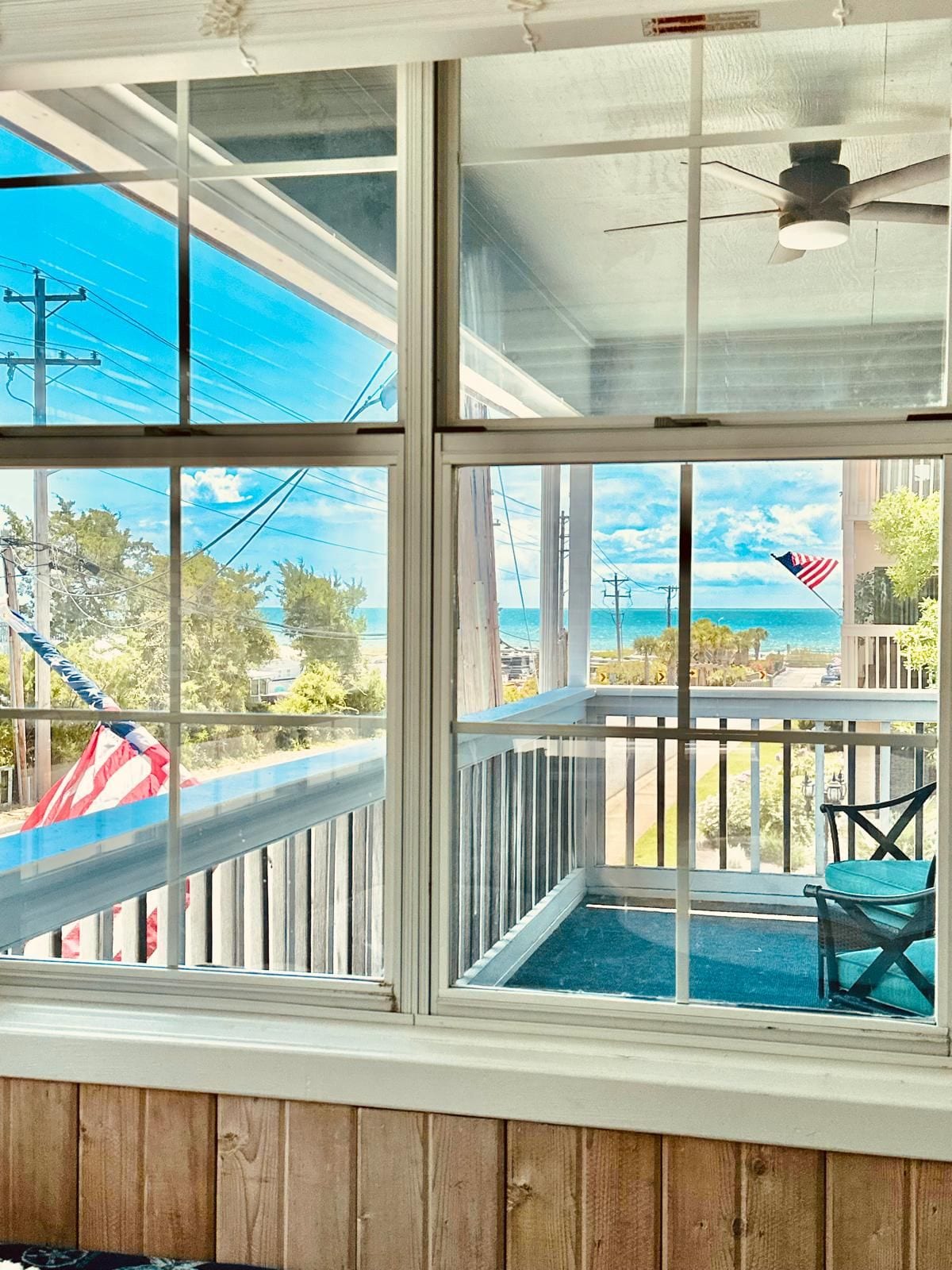A room with a large window overlooking a beach, a ceiling fan, and a view of the ocean.