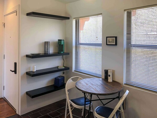A compact room with a door, a window with blinds, a round table with two chairs, and built-in shelves with various items.