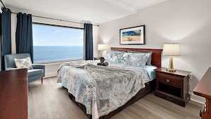 Ein Schlafzimmer mit großem Fenster, einem Bett mit gemustertem Überwurf, einem Sessel, einem Nachttisch mit Lampe und einem gerahmten Bild an der Wand.