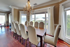 A dining area with a large wooden table and six upholstered chairs, a chandelier, and large windows with curtains. - Tybee Nights (Tybee Island)
