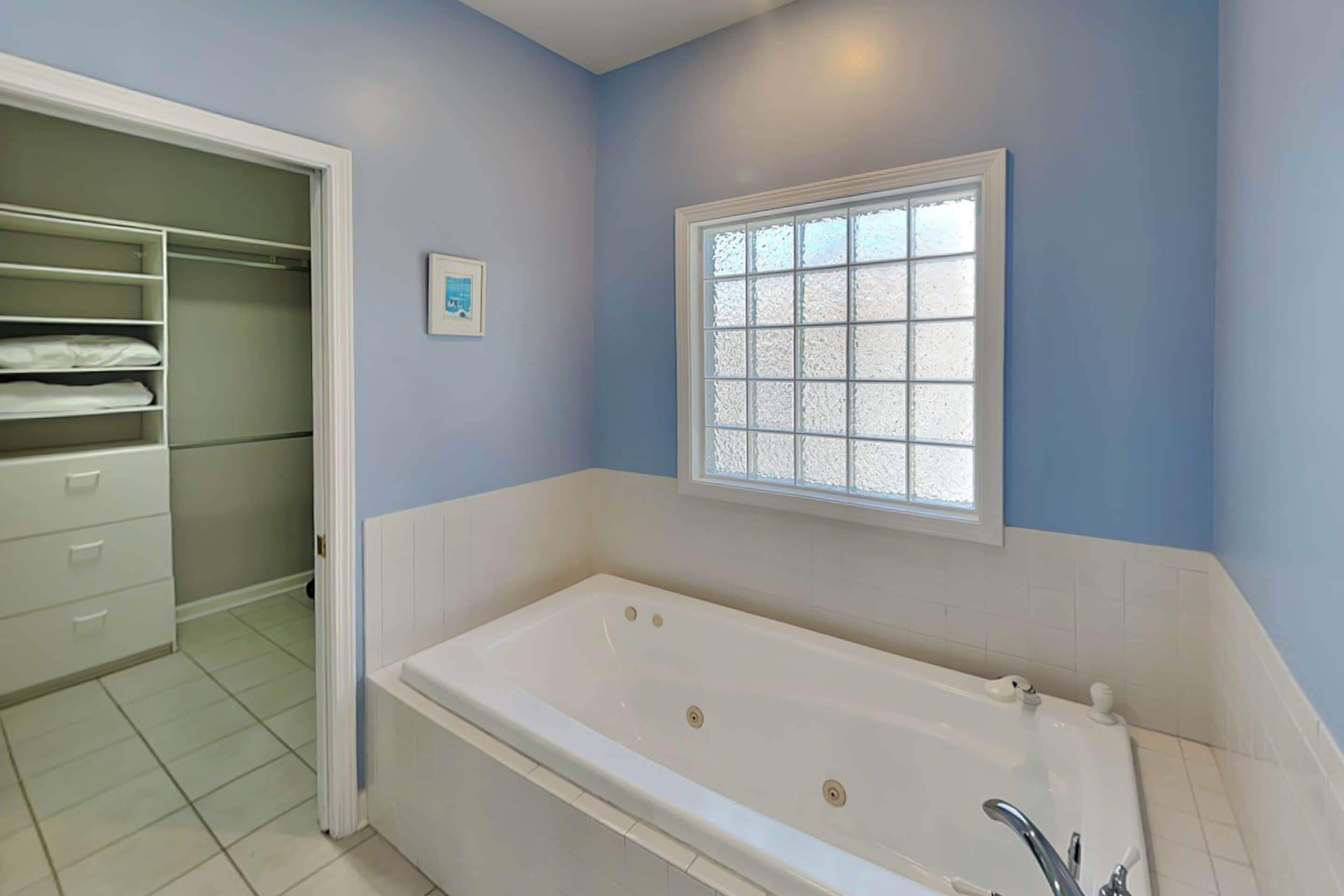A bathroom with a white bathtub, a window with frosted glass, and a walk-in closet with shelves.