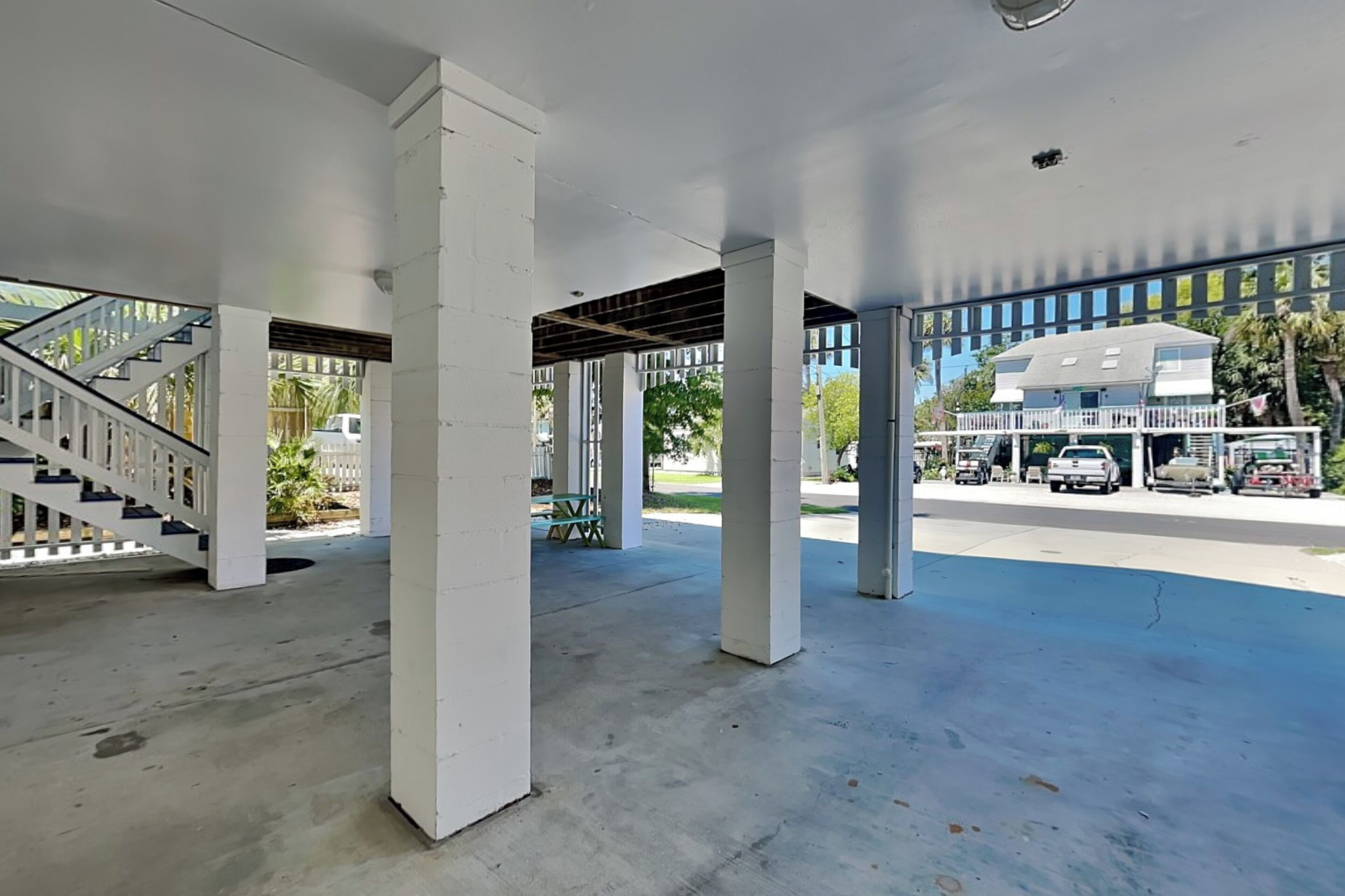 A covered outdoor area with concrete pillars, a staircase, and a view of parked vehicles and buildings in the background.