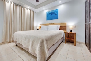 Une chambre à coucher avec un grand lit, une tête de lit en bois, deux tables de chevet avec des lampes, un ventilateur de plafond et un tableau accroché au mur.