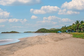 Une plage avec un ciel dégagé, un rivage sablonneux et quelques constructions, dont une tente bleue et un bâtiment avec un toit bleu.