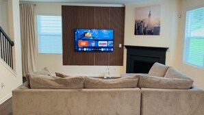 A living room with a large flat-screen TV mounted on a wooden panel wall, a beige sofa, a fireplace, and a cityscape painting. - Family Friendly and Group Stay 4BR Home, Sleeps 8 Near ATL Airport (Fairburn)
