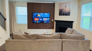 Un salon doté d’un grand téléviseur à écran plat fixé sur un mur recouvert de panneaux en bois, d’un canapé beige, d’une cheminée et d’un tableau représentant un paysage urbain.