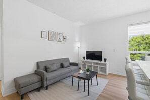 Ein modernes Wohnzimmer mit einem grauen Sofa, einem Couchtisch, einem Fernseher auf einem weißen Schrank und einem Balkon mit Grünpflanzen.