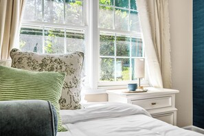 Quarto com cama, mesa de cabeceira com abajur e janela com cortinas.