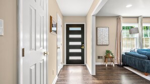 A hallway with a wooden floor, a black door, a framed picture on the wall, and a blue sofa near a window. - 5Br Lux Green Manor w/Movie Room, Fenced Yard & Sleeps 12 (Raleigh)