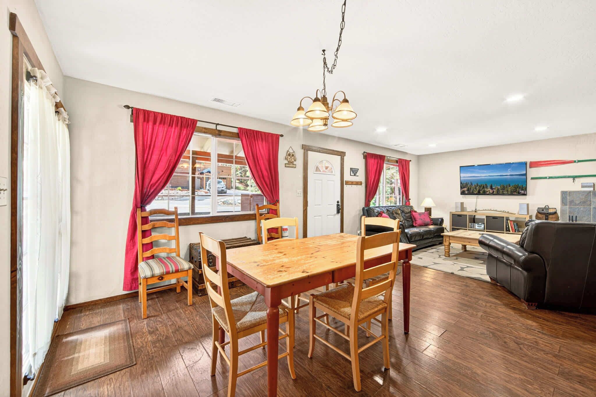 A spacious living area with a wooden dining table, red curtains, a black leather sofa, and a mounted TV.