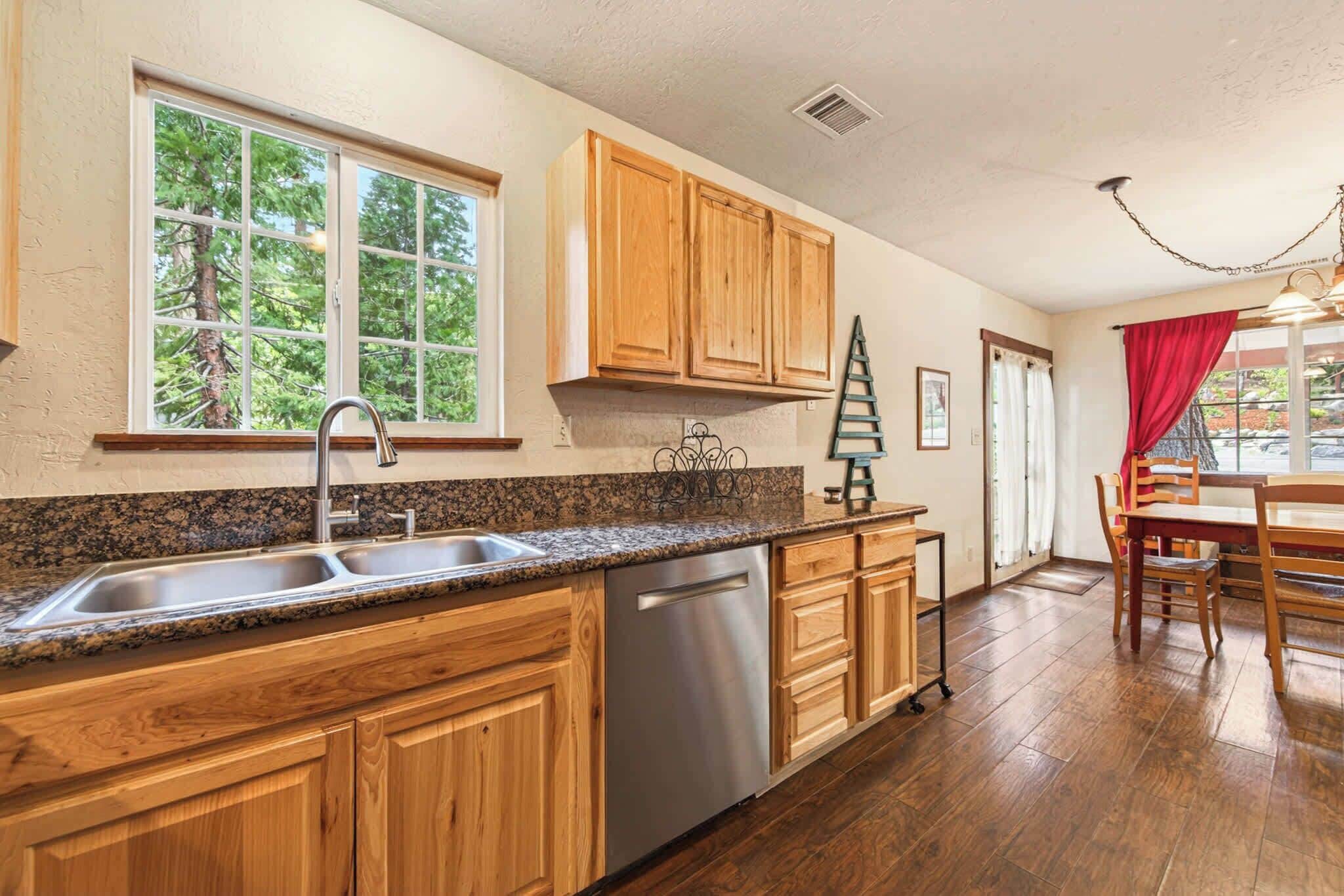 A kitchen with wooden cabinets, a stainless steel dishwasher, granite countertops, and a window with a view of trees.