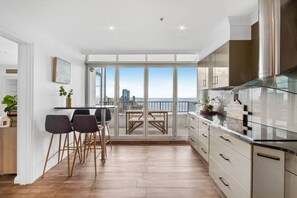 A modern kitchen with a view of the city skyline, a dining area with chairs, and a balcony with a table. - Ocean Vista Penthouse - Private Pool & BBQ, WiFi (Surfers Paradise)