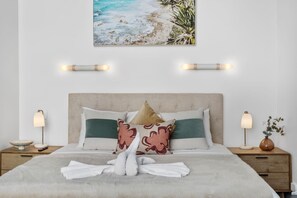 A neatly made bed with a towel arrangement, a headboard, bedside tables with lamps, and a painting of a beach scene above the bed. - Ocean Vista Penthouse - Private Pool & BBQ, WiFi (Surfers Paradise)