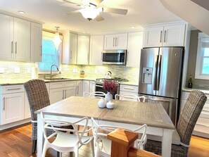 A modern kitchen with white cabinetry, stainless steel appliances, a dining table with chairs, and a ceiling fan. - Plum Island Duplex | Two Attached Townhouses (Newbury)