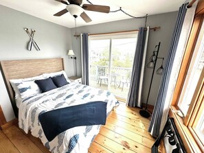 Ein Schlafzimmer mit Bett, Holzboden, Deckenventilator, einem Fenster mit Vorhängen und Blick ins Freie.