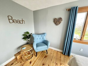 Ein Zimmer mit einem hellblauen Sessel, einem kleinen runden Tisch mit einer Pflanze, einer Wanddekoration in Herzform und einem Fenster mit blauen Vorhängen.