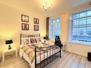 Ein Schlafzimmer mit Bett, Nachttischen, Schreibtisch, Stuhl, Fenster und Wandbildern.