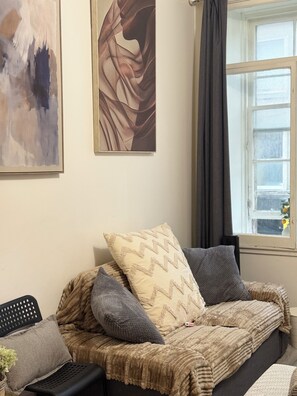 Un salon chaleureux avec un canapé, des coussins, un tableau et une fenêtre avec des rideaux.