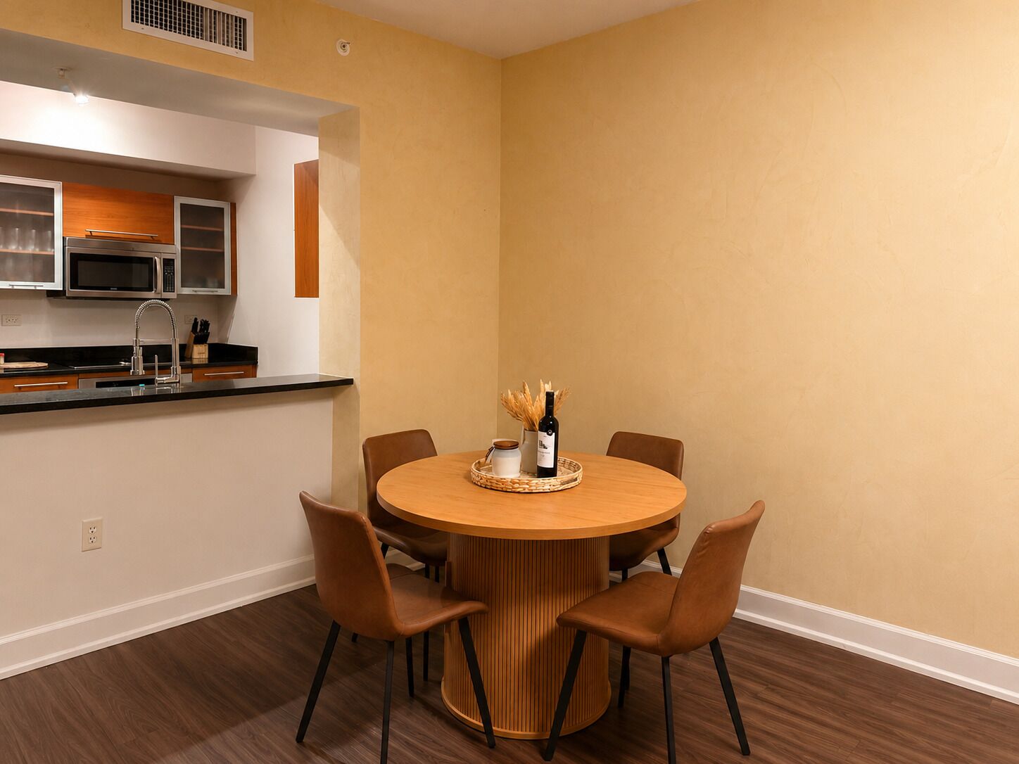 Cocina compacta con microondas, lavamanos y armarios de madera. Una mesa redonda con cuatro sillas está ubicada en el centro de la habitación.