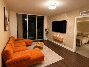 Ein modernes Wohnzimmer mit einem Ecksofa, einem Flachbildfernseher, einem kleinen Couchtisch und einem großen Fenster mit Blick auf die Stadt.