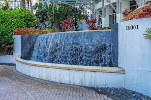 A water feature with cascading water, a concrete planter with plants, and a building with the number 18001. - Exclusive Trump Beachfront Condo (Sunny Isles Beach)