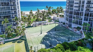 A tennis court surrounded by lush greenery and palm trees, with a view of the ocean and beach in the background. - Exclusive Trump Beachfront Condo (Sunny Isles Beach)