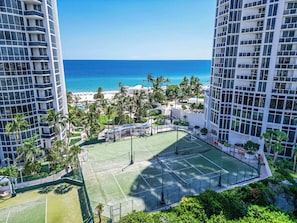 Una cancha de tenis rodeada de exuberante vegetación y palmeras, con vista al océano y a la playa.