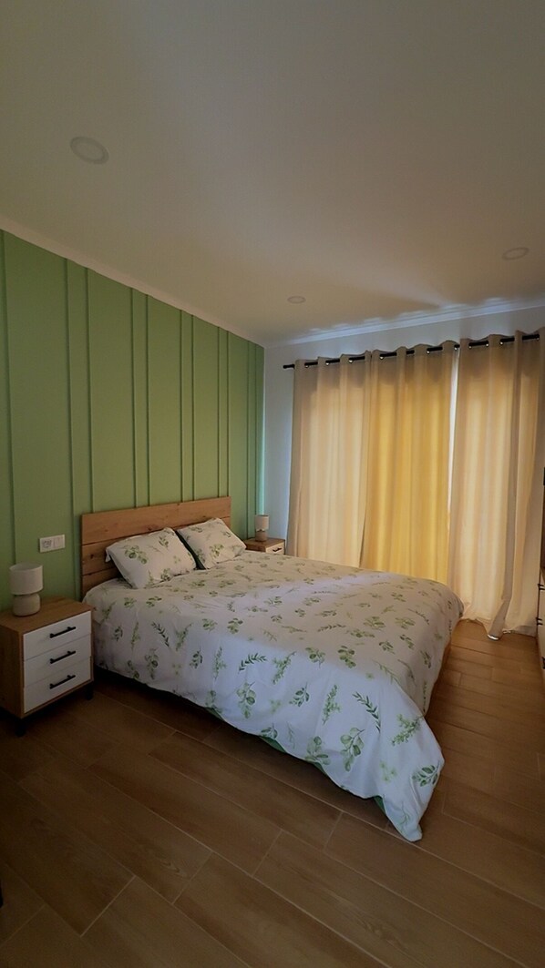 A bedroom with a wooden bed, a nightstand, a drawer unit, and a window with curtains.