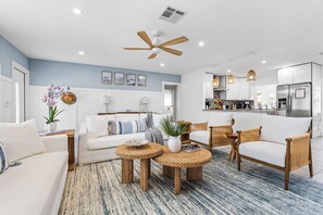 A modern living room with a sofa, two round wooden side tables, a ceiling fan, and a kitchen area with a refrigerator and pendant lights. - Backyard Oasis, Pool, Arcade & Play, Sep Deal (Delray Beach)