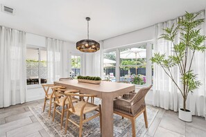 A dining room with a wooden table and chairs, a hanging light fixture, and a large window with curtains. - Backyard Oasis, Pool, Arcade & Play, Sep Deal (Delray Beach)