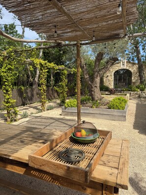 Eine hölzerne Terrasse im Freien mit Grill und einer Fruchtschale, umgeben von einem rustikalen Steinhaus und viel Grün.