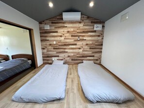 A room with two beds, wooden wall paneling, an air conditioning unit, and a visible electrical outlet. - Hokkaido Hideaway: Bonfire & BBQ (Toyoura)